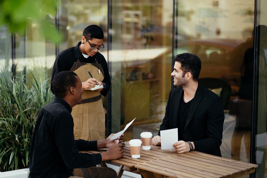Two people at an outdoor café table with a server taking an order, talking face to face
